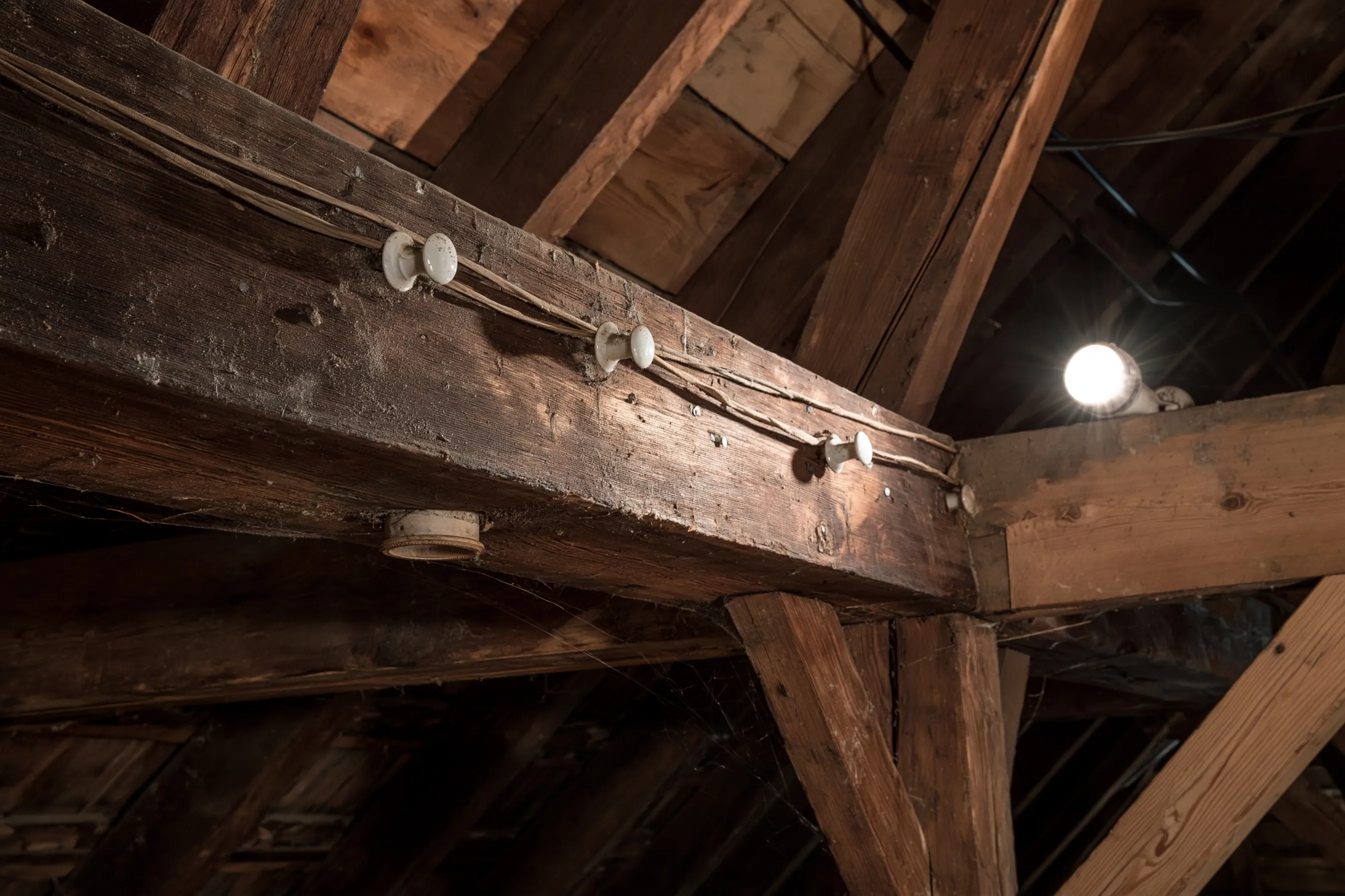 Knob-and-tube wires on white porcelain knobs running along dark attic beams, with a bare bulb lit nearby